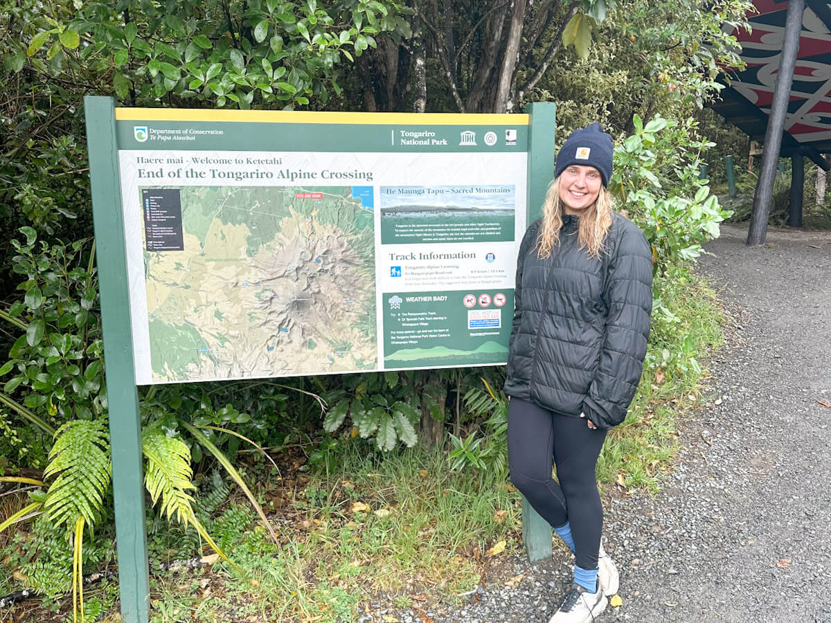 Charlie is standing next to an official Tongariro National Park sign at the end of the Tongariro Alpine Crossing, providing track information and a map of the famous New Zealand hike
