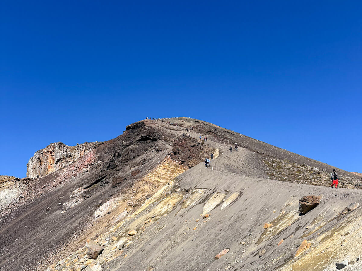 Hikers ascending a steep, rocky ridge towards a summit on the Tongariro Alpine Crossing, showing the challenging but rewarding climb with a clear blue sky in New Zealand.