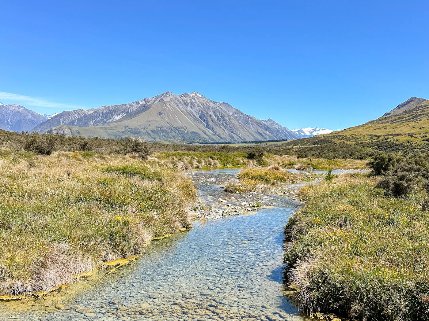 A clear stream flows through a grassy, tussock-covered valley with the majestic Mount Sunday in the background, a famous Lord of the Rings filming location in the Canterbury High Country of New Zealand's South Island.