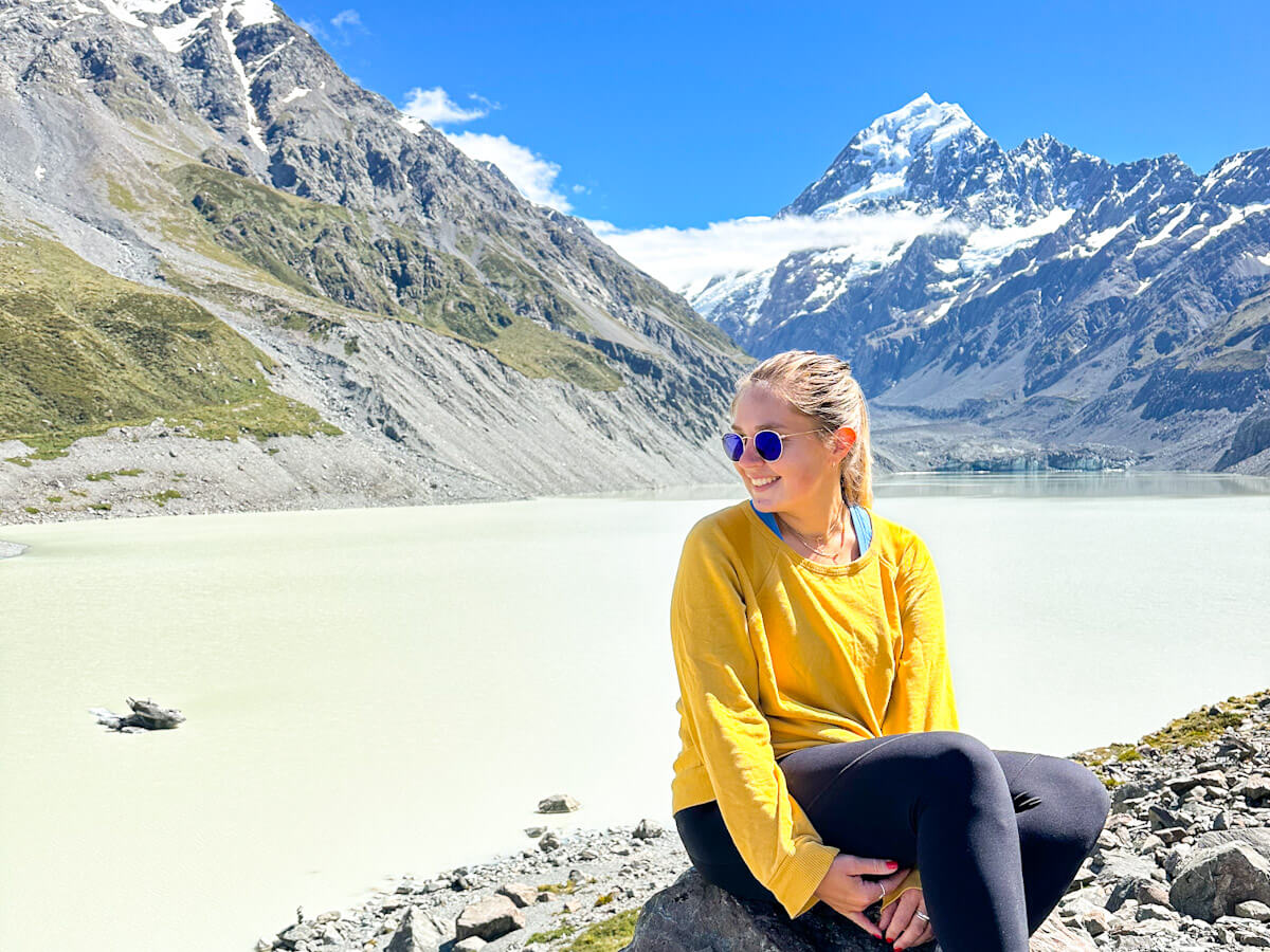 Charlie is wearing a yellow jumper, black leggings, and blue sunglasses looking to the side and smiling. She is sitting on a rock with a turquoise lake behind her, and the peak of Mount Cook in the background at the end of the Hooker Valley Trail