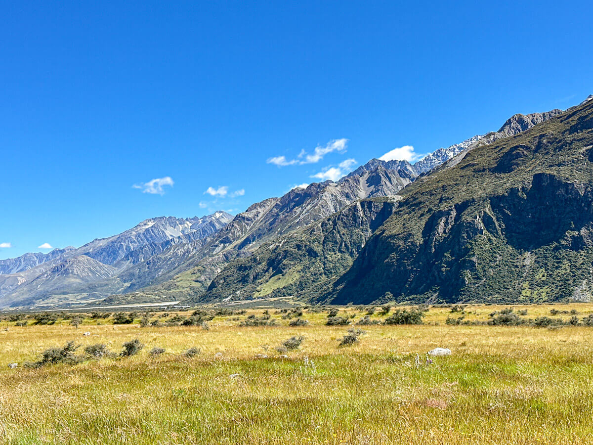 A wide shot of the vast golden fields of Aoraki/Mount Cook National Park, with a long mountain range stretching across the horizon under a clear blue sky. Showing the spot where the Lord of the Rings filming location for Minas Tirith sat