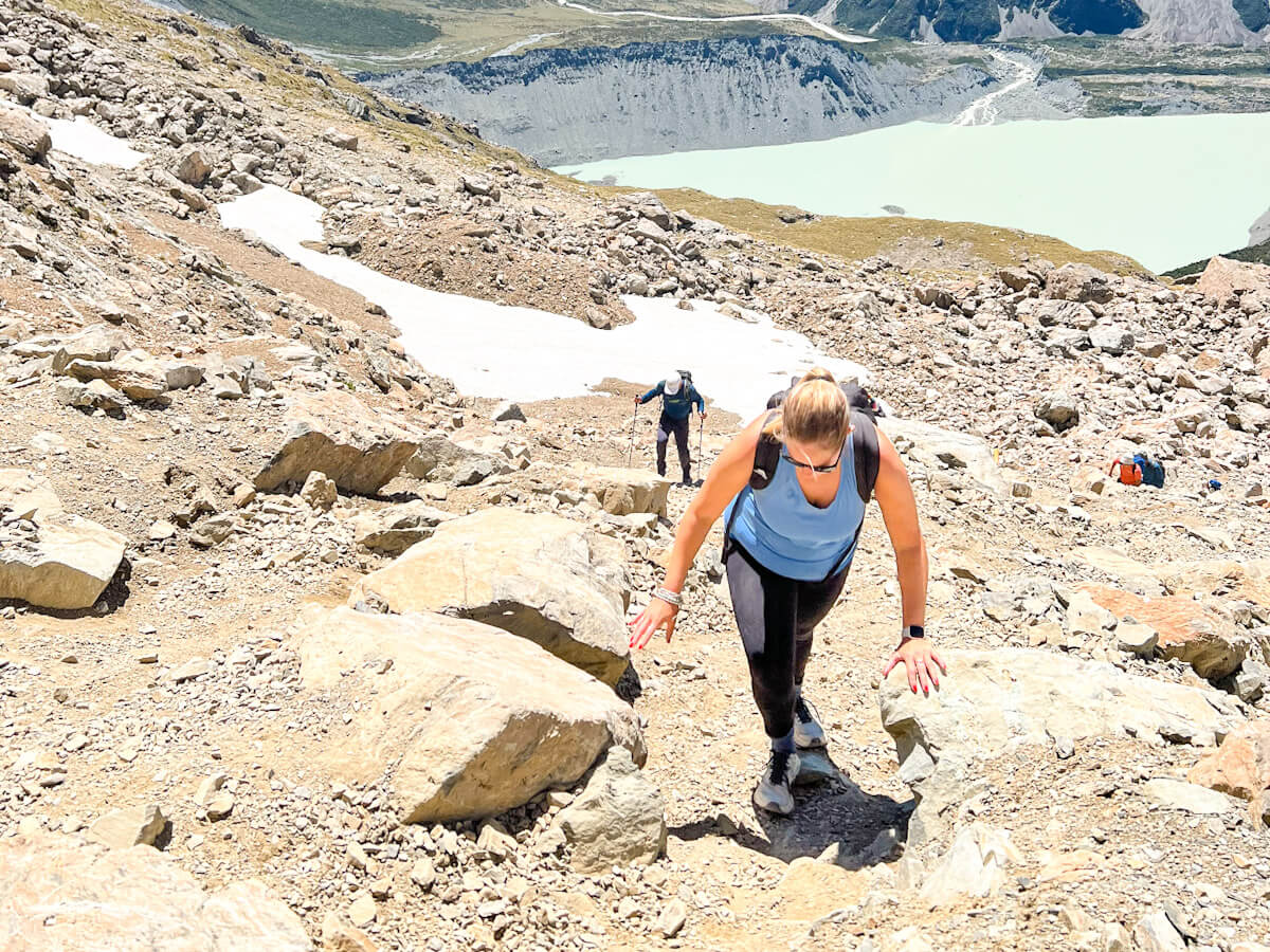 Charlie climbs a rocky trail on the Mueller Hut Track with a panoramic view of the glacial Mueller Lake and the surrounding Mount Cook mountains behind them.
