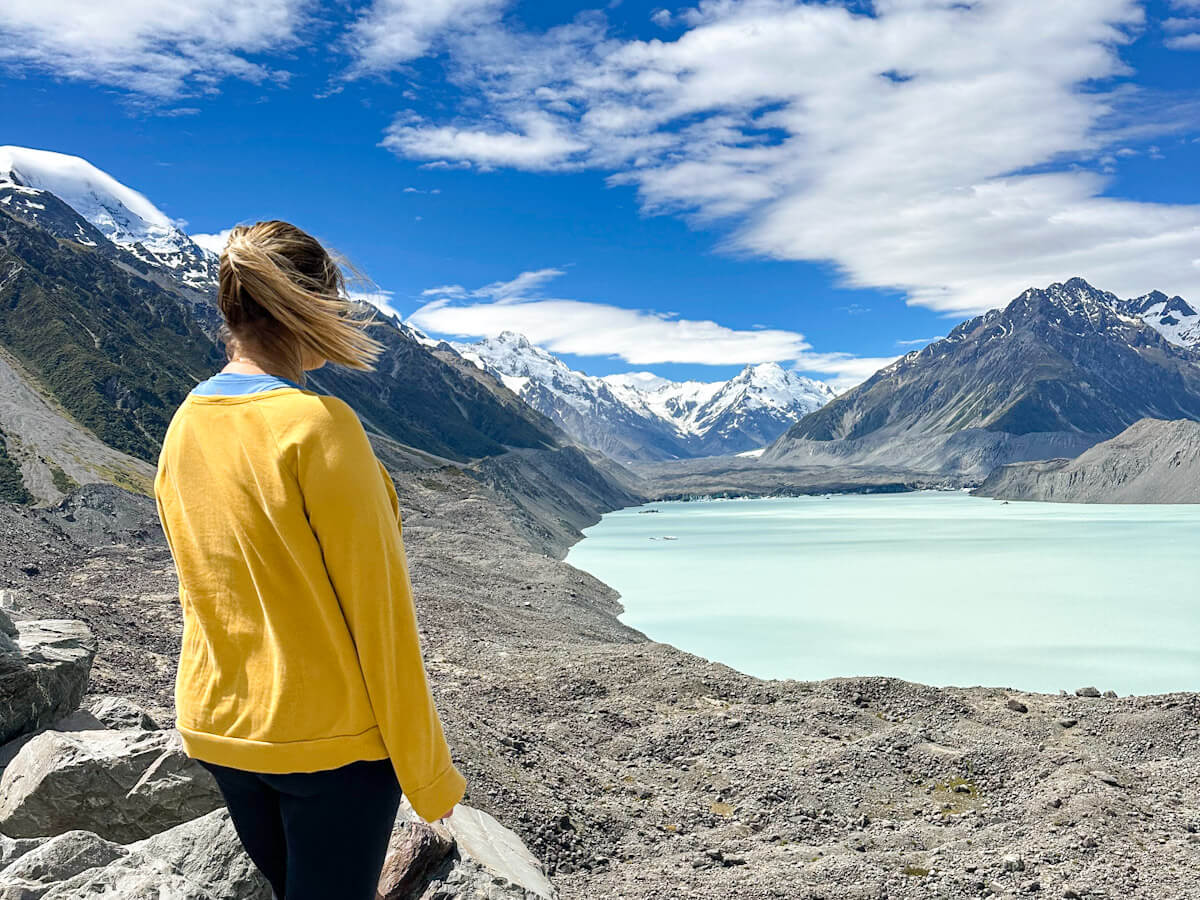 Charlie stands on a rocky viewpoint looking out at the milky turquoise waters of the Tasman Lake and the immense Tasman Glacier flowing from the snow-covered mountains.