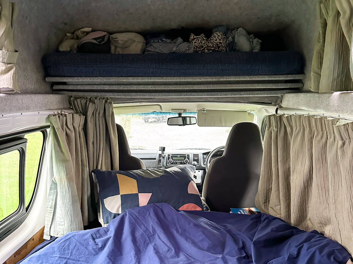 Interior shot of the campervan showing the bed with blue sheets and patterned pillows, and a raised storage area above the cab, set up for solo travel in New Zealand.