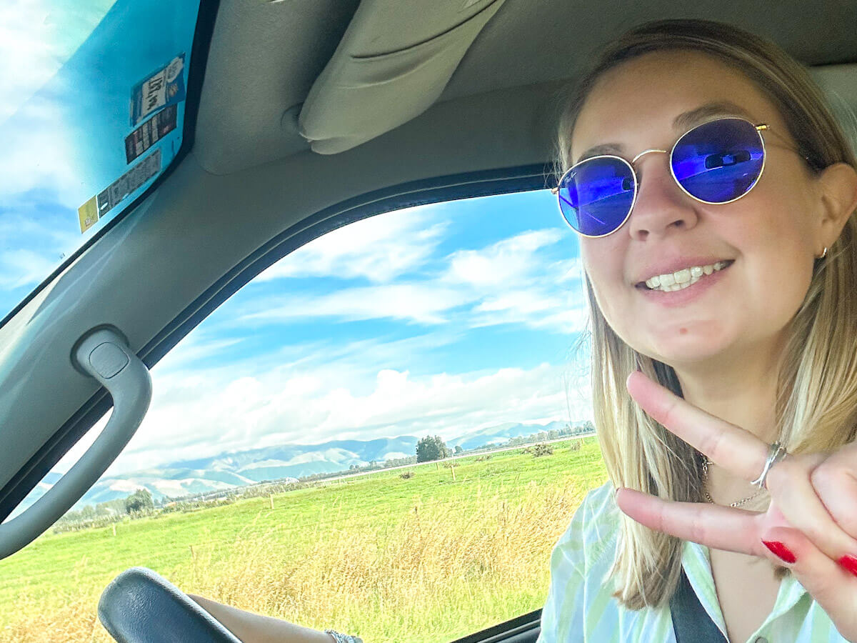Charlie is smiling and making a peace sign while driving the campervan on a sunny day with green fields and distant hills, enjoying a solo travel journey.