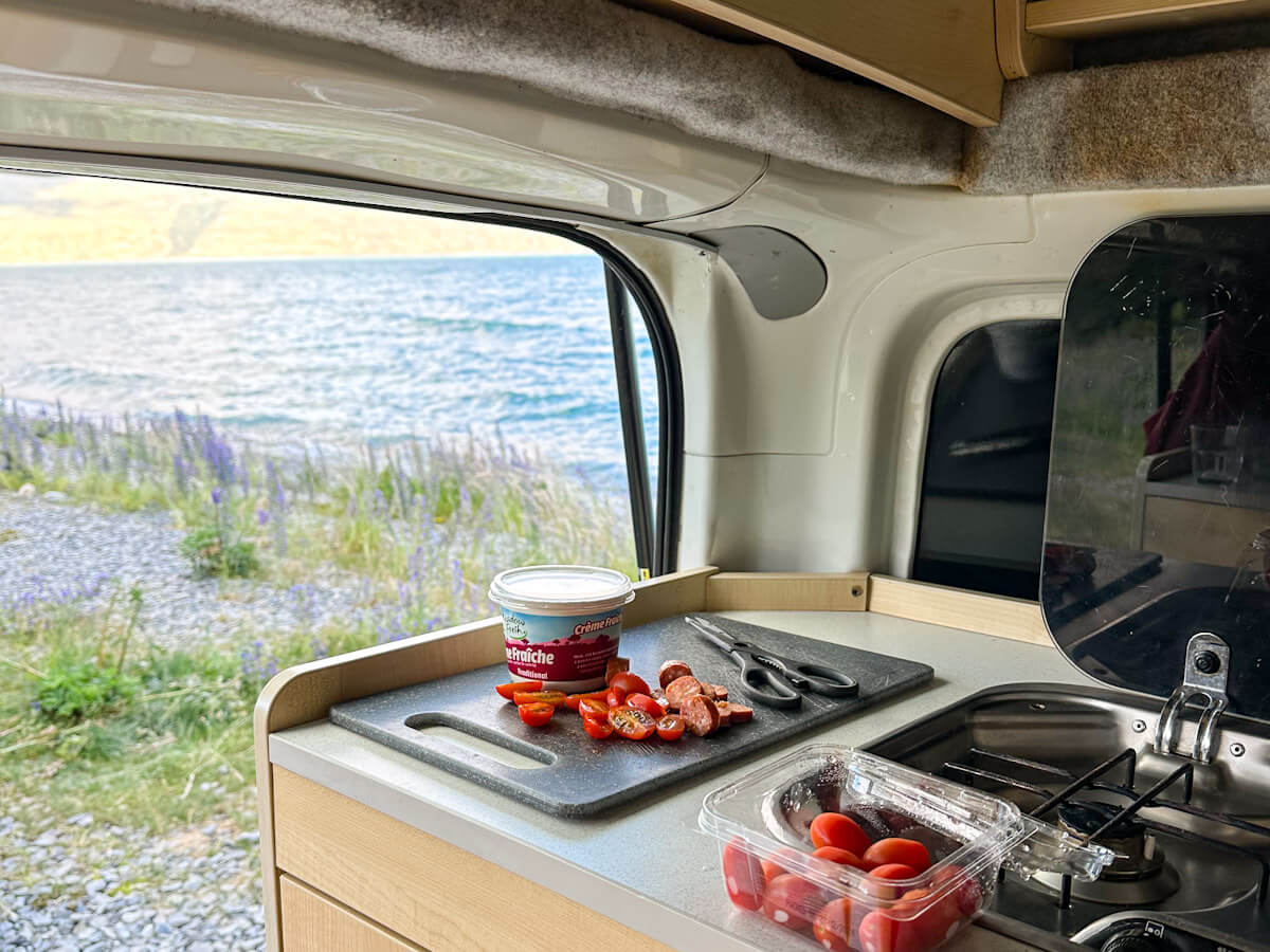 Interior shot of a campervan kitchen counter with chopped tomatoes and sausage on a cutting board, with a view of a blue lake and purple lupins outside, highlighting solo travel cooking.