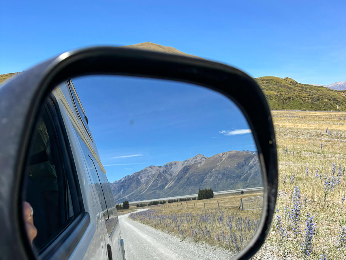 View in the campervan's side mirror of the gravel road leading to Mount Sunday (Edoras) with rugged mountains and yellow fields of grass, highlighting the scenic drive during solo travel in New Zealand.