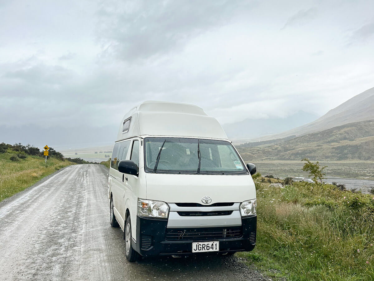 A white high-top campervan is on a muddy, misty gravel road in a scenic New Zealand valley, on the road to Mount Sunday, symbolising the freedom of solo travel.