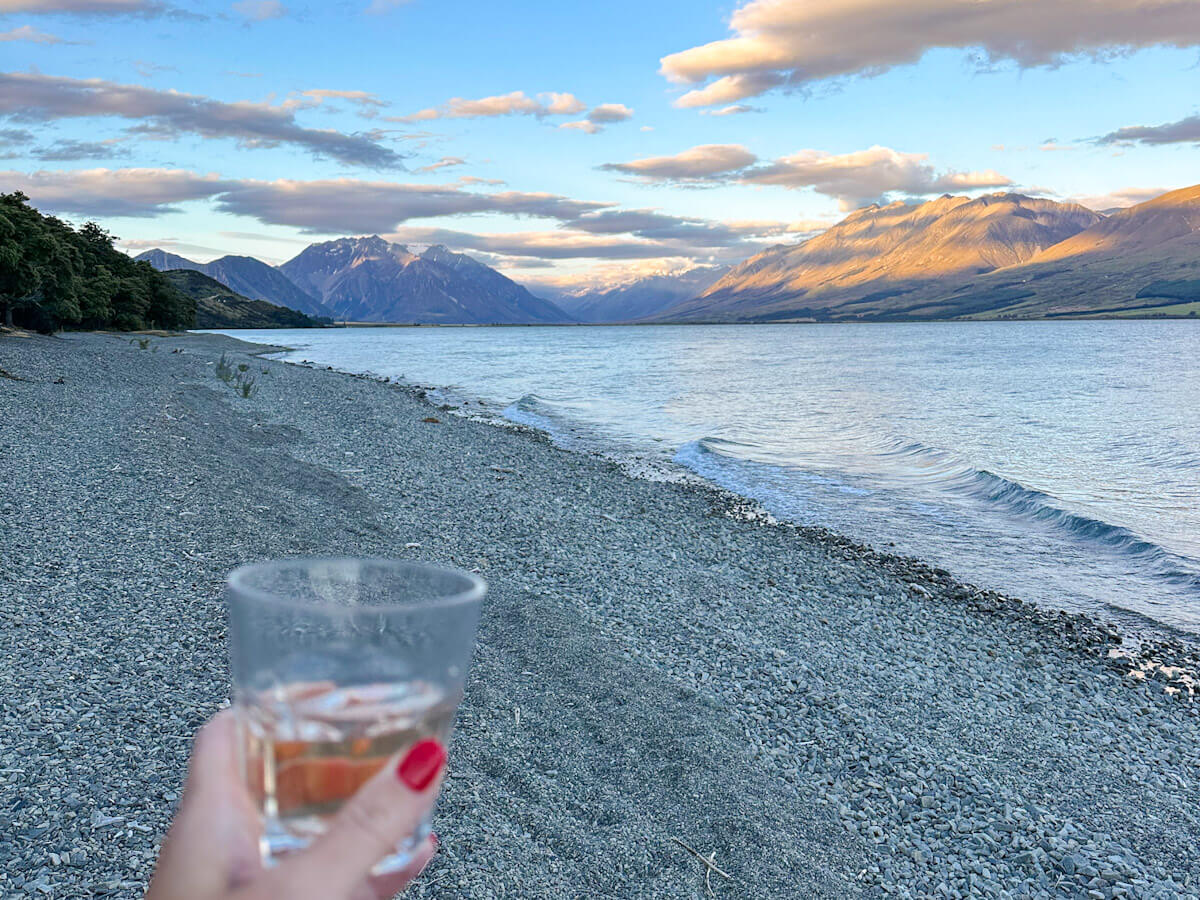 A hand with red-painted nails holding a glass of rosรฉ wine on a shingle beach, with the vast lake ohau and sun-kissed mountains in the background, a serene moment of solo travel in New Zealand.
