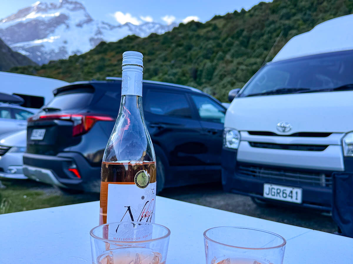 A bottle of Marlborough Rosรฉ wine and two glasses on an outdoor table, with the campervan (JGR641) and a snow-capped mountain peak in the background, enjoying a solo travel evening.