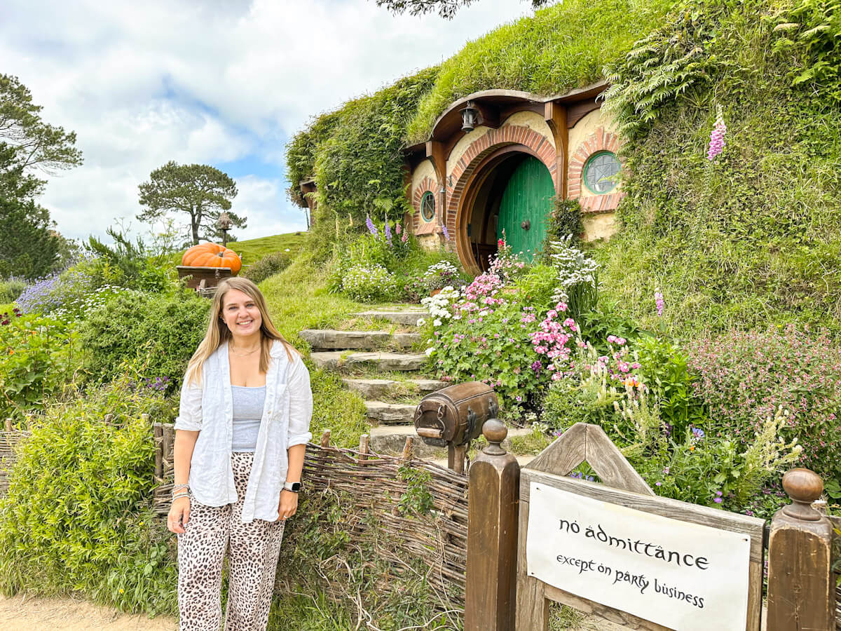 Charlie stands in front of the hobbit hole Bag End, with a green door and an archway of stone steps, featuring a sign that says, "No admittance except on party business," during her visit to Hobbiton.