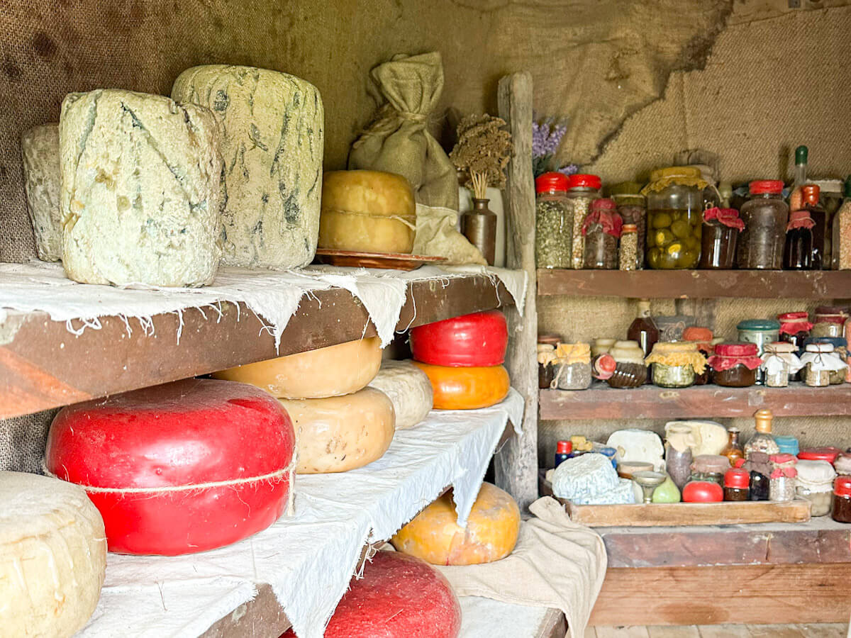 A detail shot of a hobbit hole pantry at Hobbiton displaying large wheels of red and natural-coloured cheese, alongside jars of preserved goods and dried herbs.
