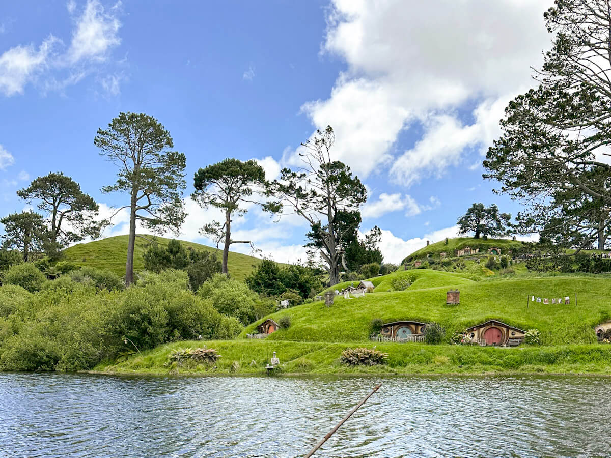 A scenic view across the water toward the Hobbiton Movie Set, featuring several colorful hobbit holes dug into the lush green banks under a bright, cloudy sky.