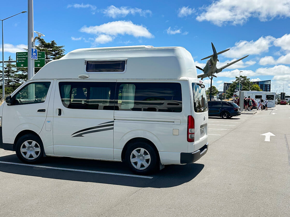 Side view of the white high-top campervan parked in a lot, with a historic airplane on display in the background, likely at a New Zealand airport, marking the start of a solo travel adventure.