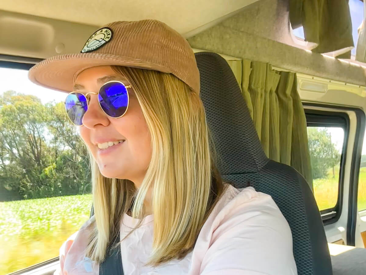 Charlie is wearing a corduroy cap and blue mirrored sunglasses, smiling while driving the campervan through a green landscape, embodying solo travel in New Zealand