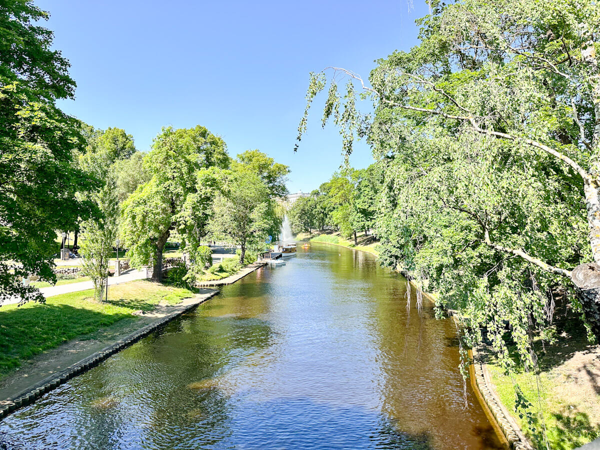 What to do in Riga: A view down the peaceful Riga City Canal (Pilsētas kanāls), surrounded by lush green trees, with a small canal boat visible in the distance.