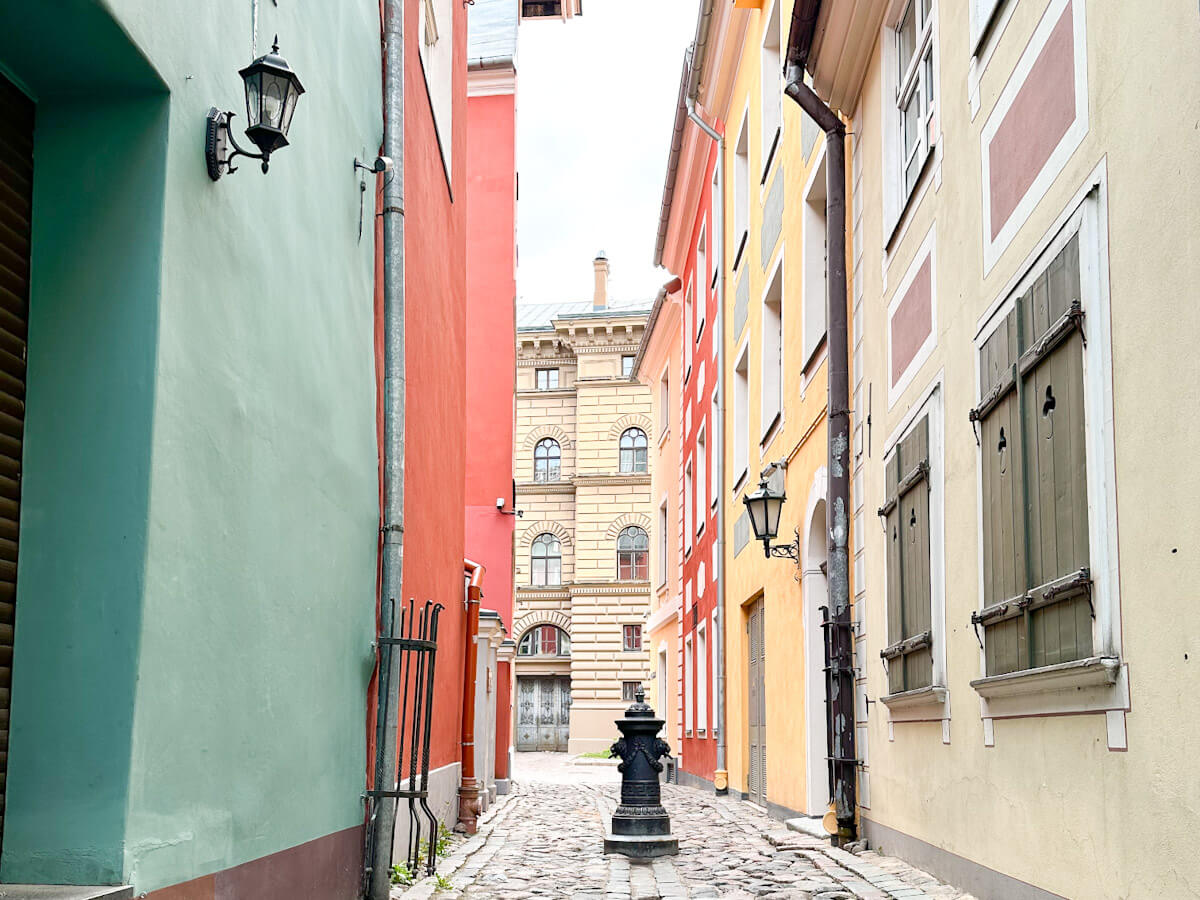 What to do in Riga: A view down a narrow cobblestone alleyway in Old Town, flanked by brightly painted red and mint-green buildings, with an old water pump in the center.