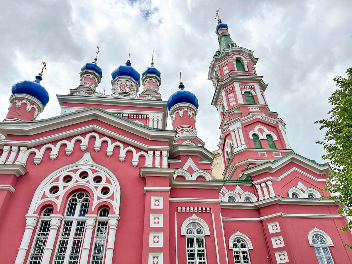 What to do in Riga: An architectural view of the vibrant pink and white facade and bright blue onion domes of the Russian Orthodox Church of Our Lady of Sorrows.