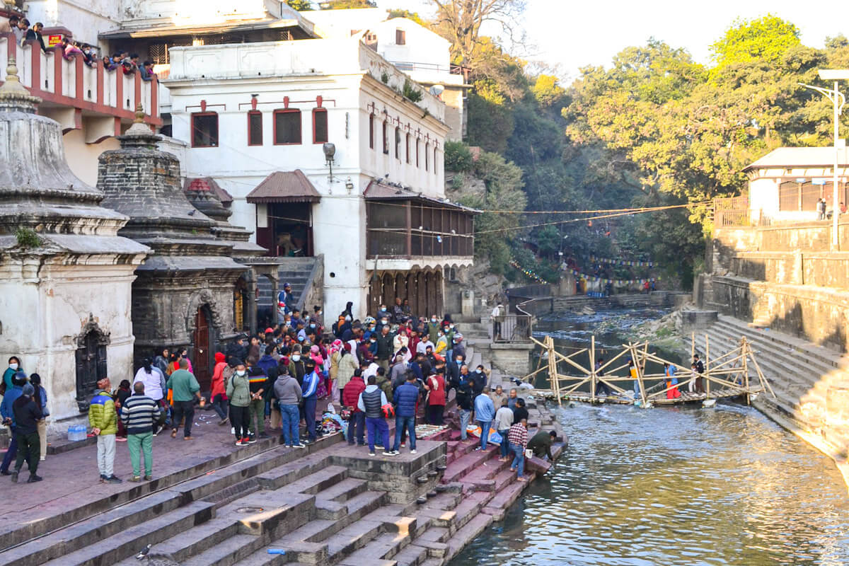 A view of the Bagmati River with a crowd of people gathered on the stone steps (ghats) next to a white building, at the Pashupatinath Temple area, highlighting the cultural places to visit in Kathmandu