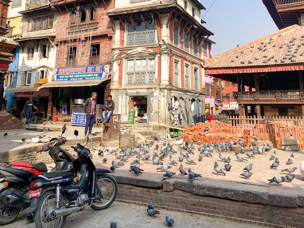 A sun-drenched, historic square in the Kathmandu Valley, showing traditional multi-story Newari brick buildings, parked motorbikes, and many pigeons gathering on the ground. Exploring the ancient squares is one of the best places to visit in Kathmandu.