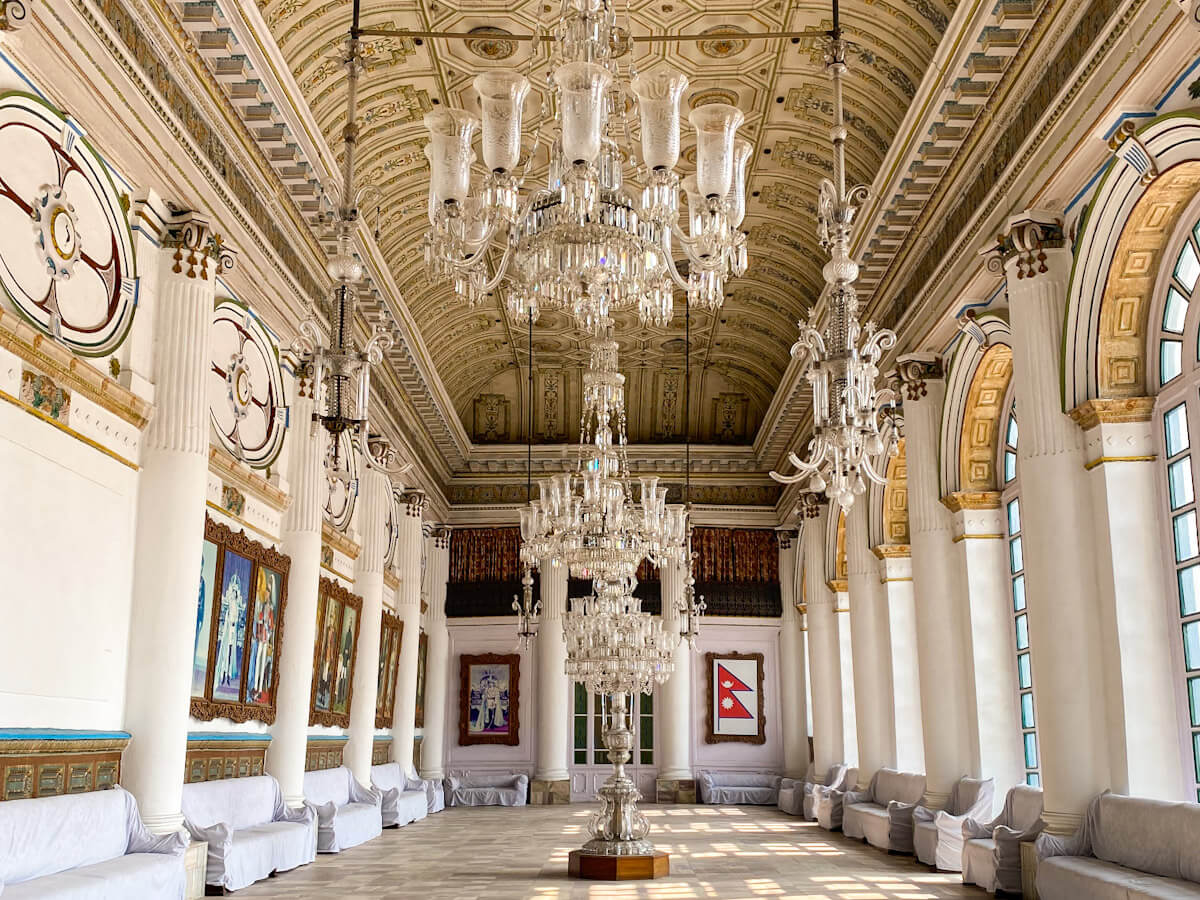 The grand interior hall of Hanuman Dhoka in Kathmandu, featuring ornate crystal chandeliers, tall white columns, and paintings. A beautiful example of the historical places to visit in Kathmandu.