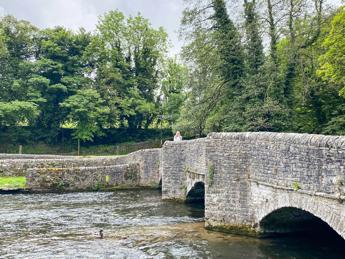 The historic stone Sheepwash Bridge crossing a clear river in Ashford-in-the-Water, one of the best villages in the Peak District, with a duck swimming in the water.