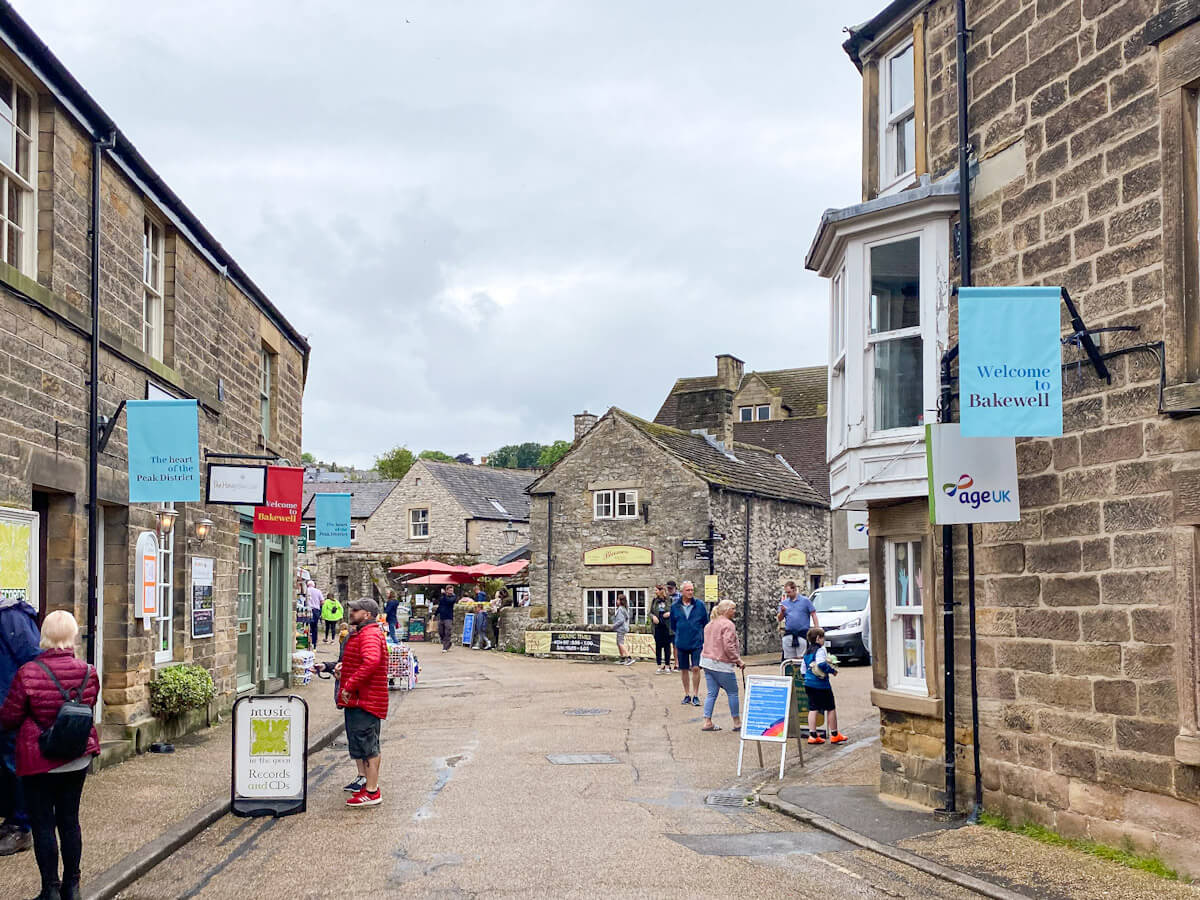 The bustling town center of Bakewell, one of the best villages in the Peak District, showing pedestrians walking past local shops and "Welcome to Bakewell" flags.