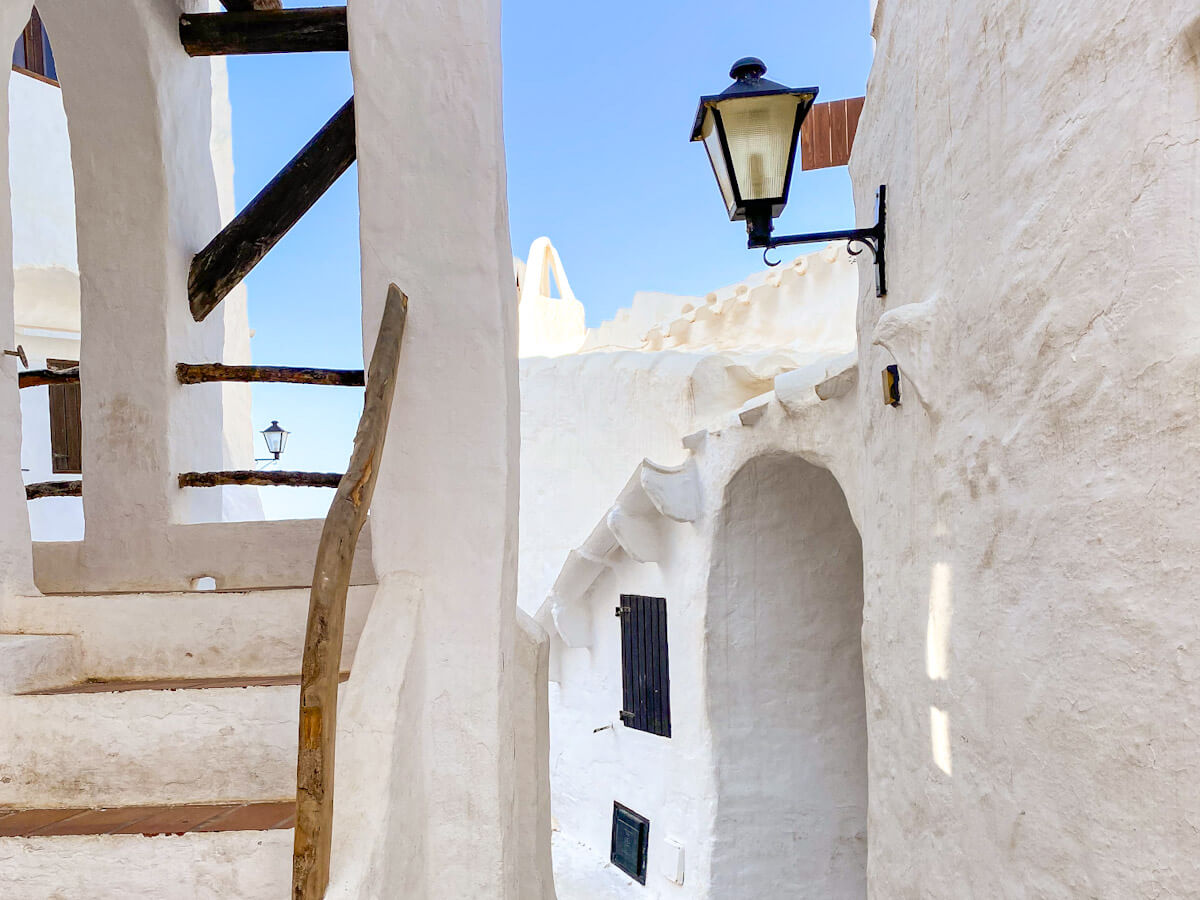 A bright, sun-drenched narrow alleyway in Binibeca Vell, featuring smooth white-plastered walls, a traditional black street lamp, and rustic wooden staircase railings.