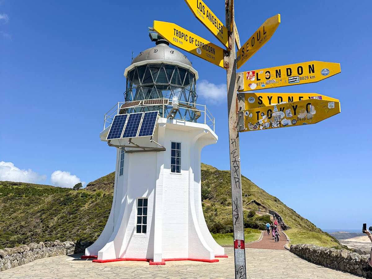 The white Cape Reinga lighthouse and a yellow signpost pointing to global cities, a highlight of a Northland day trip for those searching for what to do in Paihia.