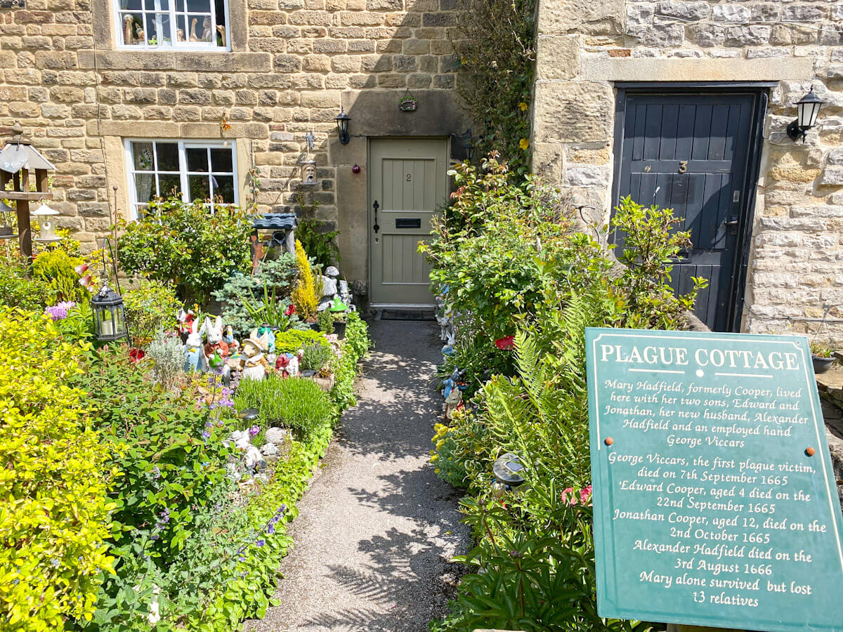 The historic "Plague Cottage" in Eyam, one of the best villages in the Peak District, featuring an informative green sign in front of a stone cottage with a garden full of flowers and decorative ornaments.