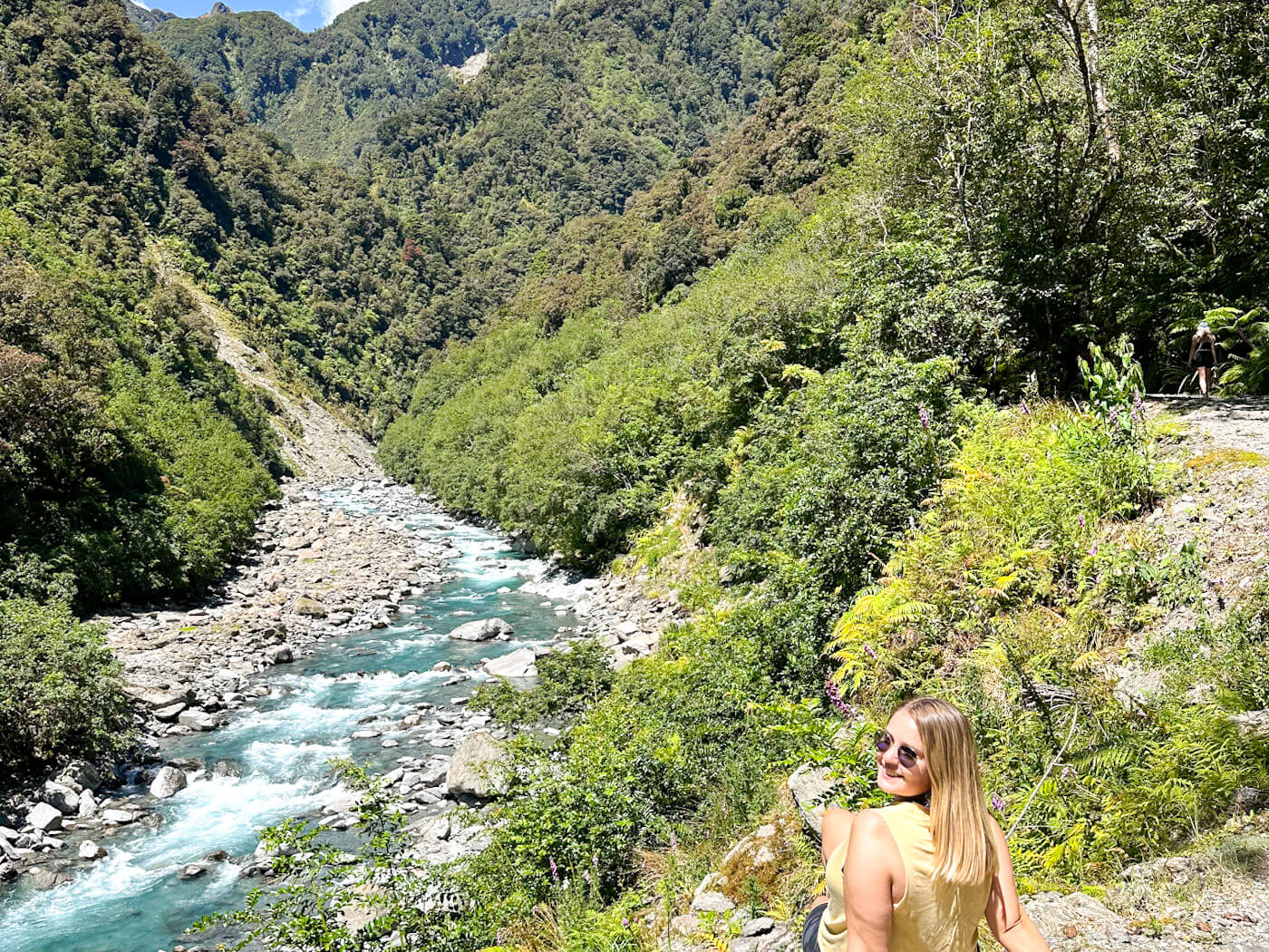 Charlie is wearing a yellow top abnd overlooking turquoise Waiho River flowing through lush green valley near Franz Josef, one of the scenic things to do in Franz Josef, New Zealand