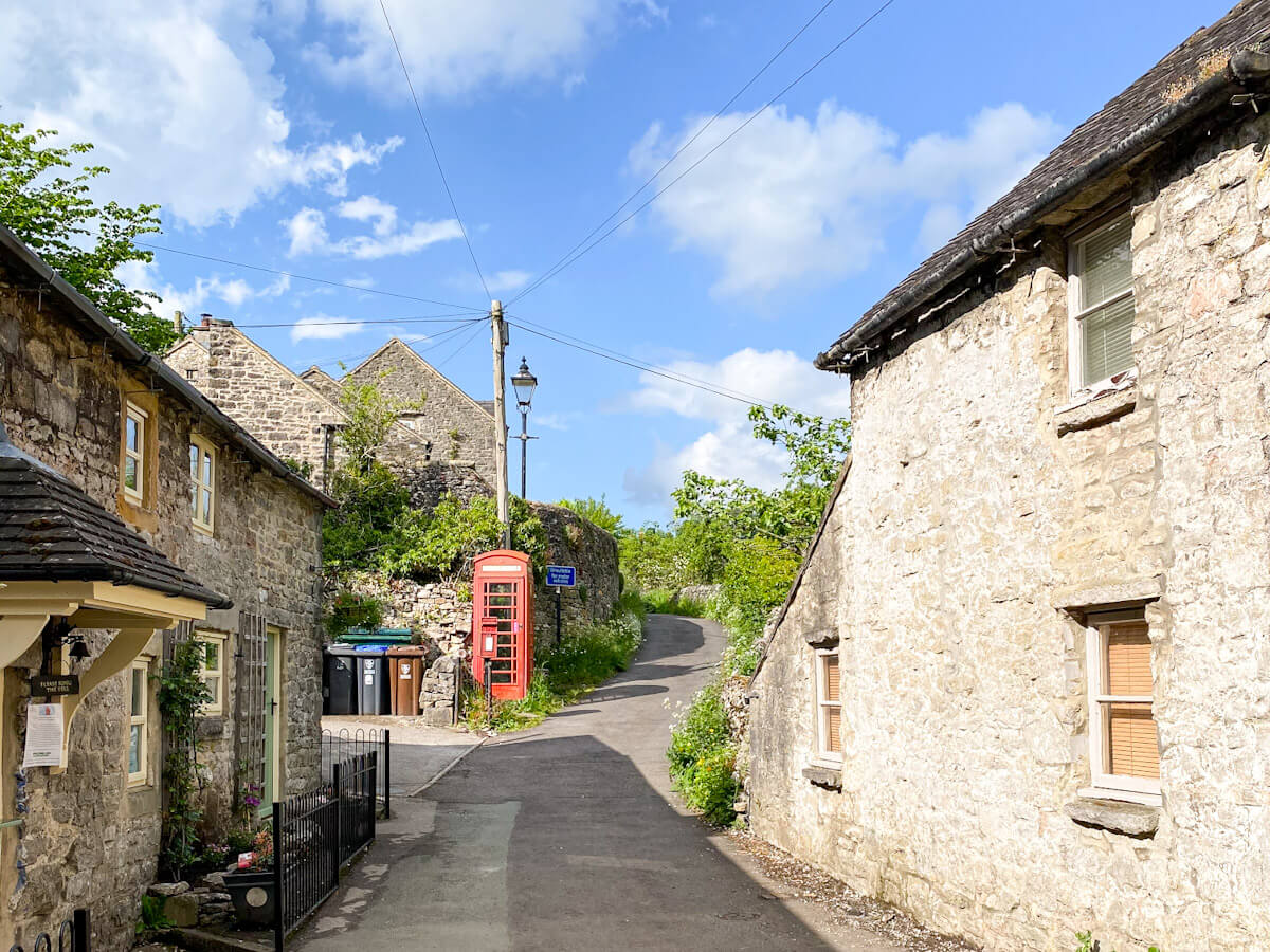 A quiet, narrow road through the tiny hamlet of Milldale, one of the best villages in the Peak District, featuring a classic red British telephone box next to historic stone buildings.