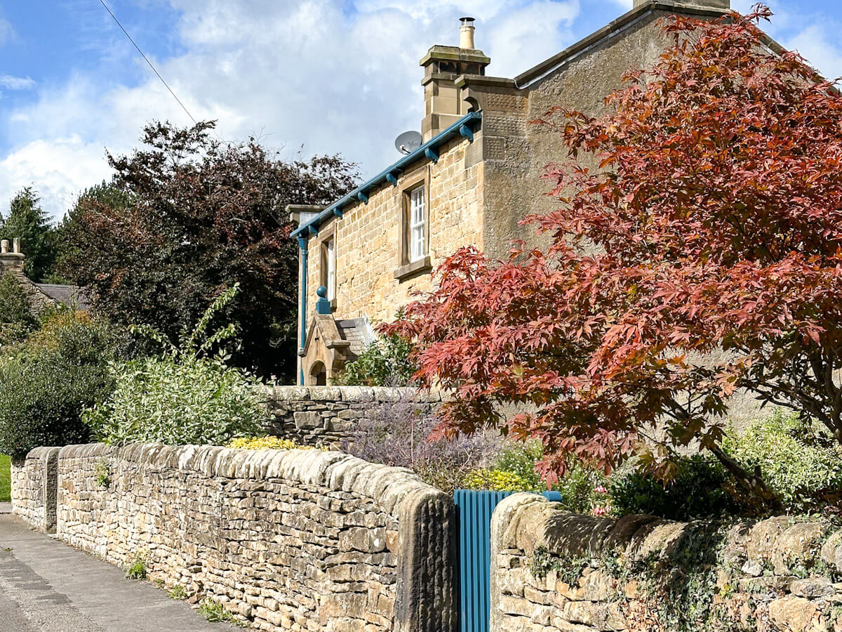 A charming stone cottage in Pilsley, one of the best villages in the Peak District, featuring a low dry stone wall and a vibrant red-leaved tree in a well-kept garden.