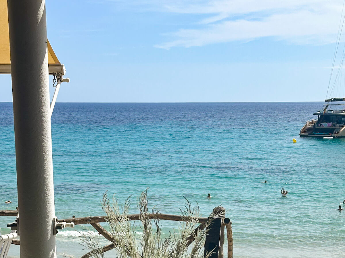 Looking out over the expansive turquoise and deep blue Mediterranean Sea at Santo Tomรกs, a highlight among the best beaches in Menorca, with a luxury yacht anchored in the distance and swimmers in the water.