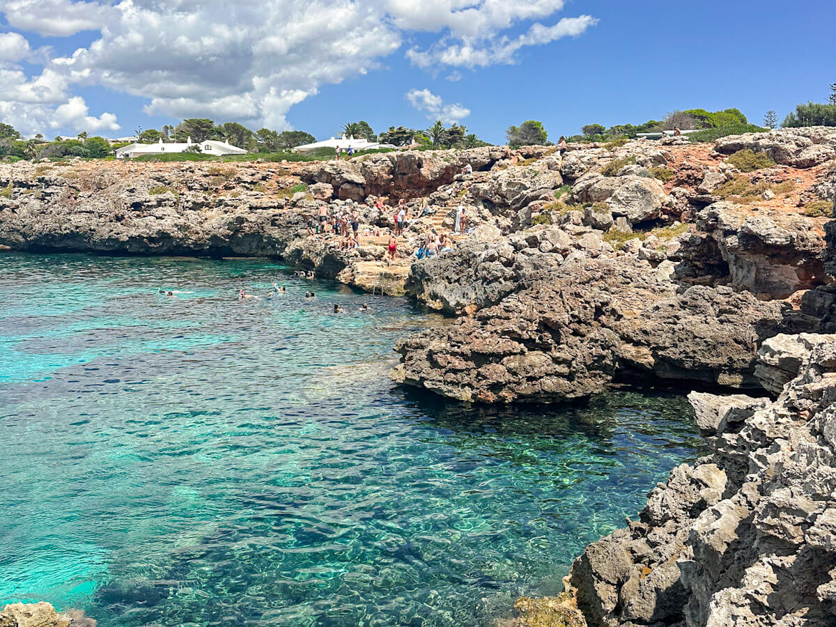 A rocky swimming area at Ses Olles with clear, bright turquoise water where people are swimming and sunbathing on the craggy limestone cliffs.