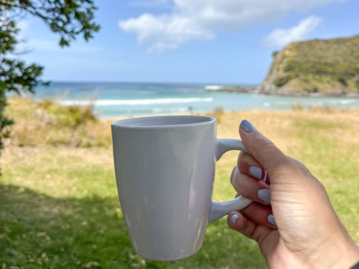 A person holding a white mug with a blurred view of the turquoise beach and headland of Tapotupotu Bay in the background, showing relaxing things for what to do in Paihia.