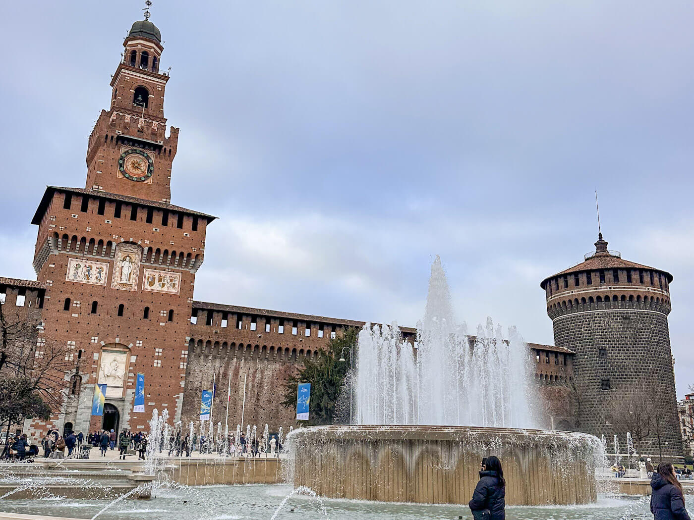 A wide shot of the red-brick Sforza Castle (Castello Sforzesco) featuring the iconic Filarete Tower and a round stone turret, with a large, tiered water fountain spraying white mist into the air in the foreground.