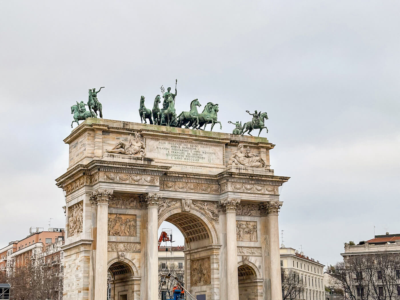 A medium shot of the top portion of the Arco della Pace, showcasing the intricate neoclassical stone reliefs and the bronze "Sestiga della Pace" (chariot of peace) statue featuring six horses and a goddess against a grey, cloudy sky.
