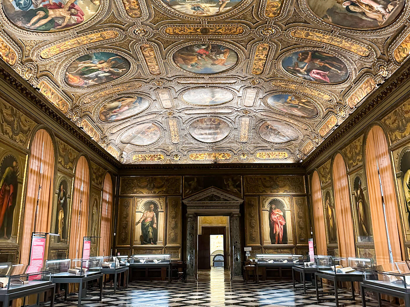 An opulent hall in museo Correr with a magnificent gold-leaf ceiling and classical paintings, featuring checkered marble floors and glass exhibit cases.