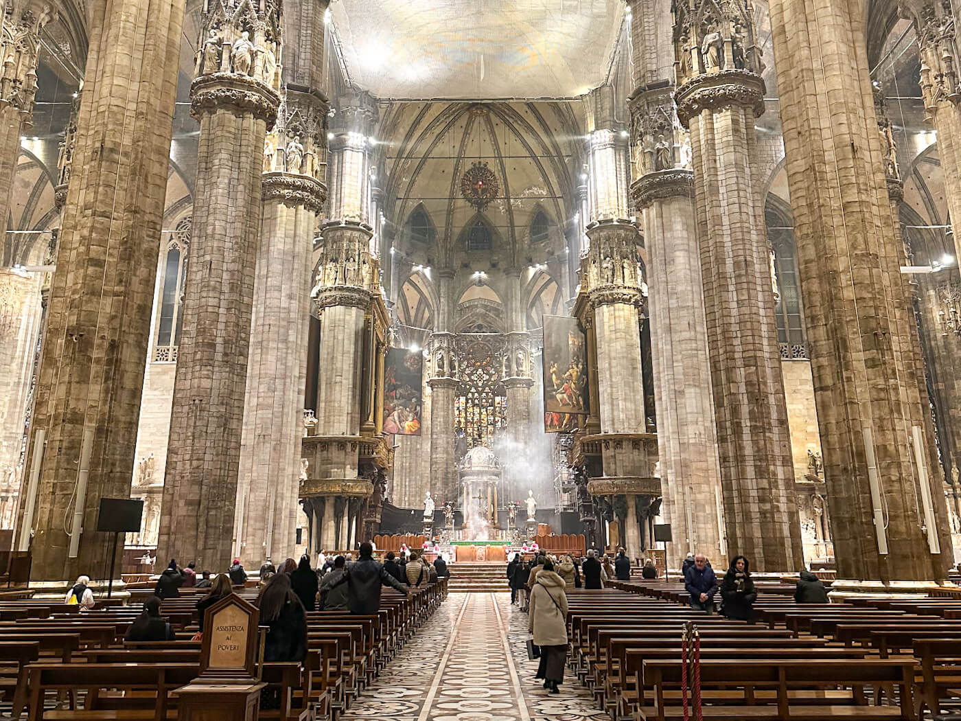 The grand interior of the Milan Cathedral (Duomo), featuring massive stone columns decorated with statues, rows of wooden pews filled with visitors, and a distant altar illuminated by soft light and colorful stained-glass windows.
