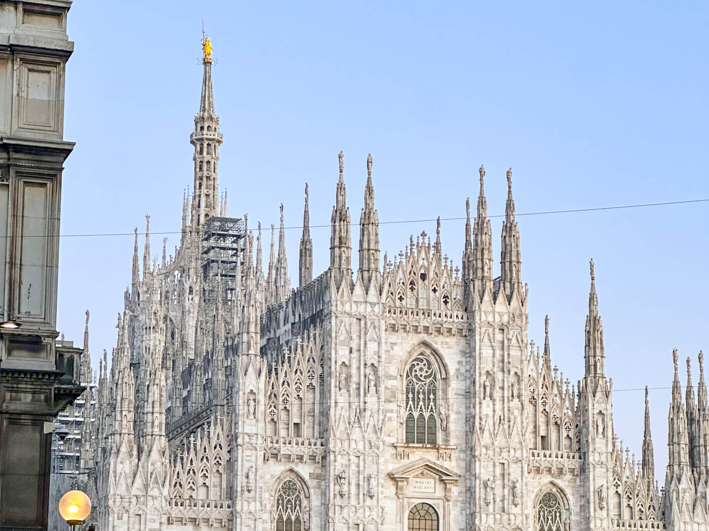 A medium shot of the upper facade of the Milan Cathedral, highlighting the detailed white marble spires and statues, with the iconic golden "Madonnina" statue perched on the tallest central spire against a pale blue sky.