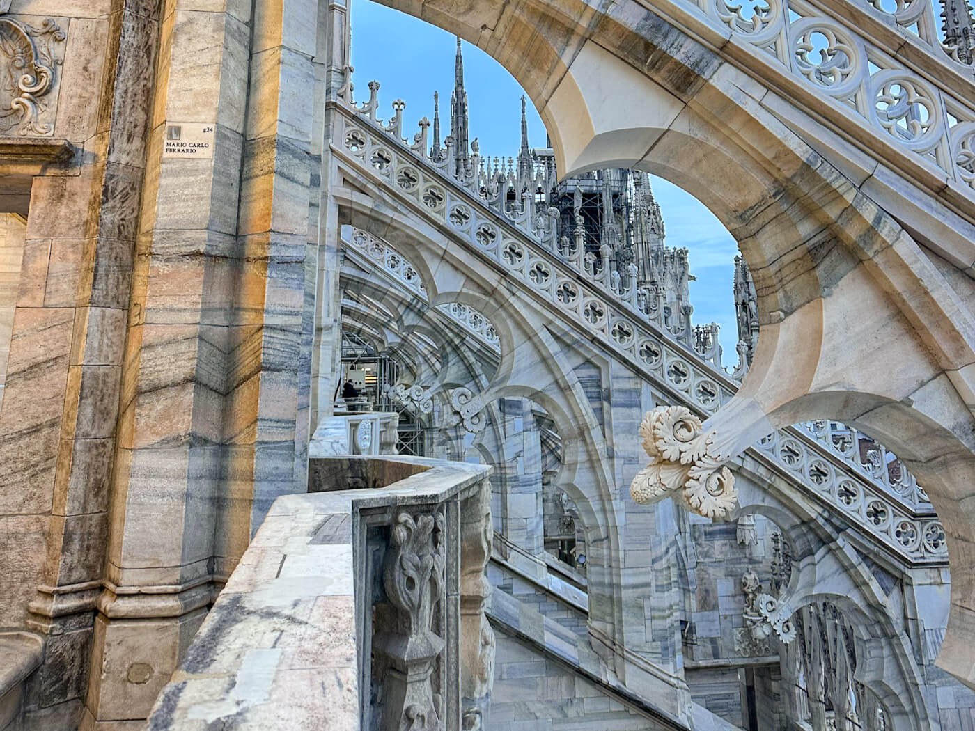 A detailed architectural shot of the Duomo di Milano's rooftop terraces, showcasing the flying buttresses, ornate marble carvings, and repetitive arched stone patterns against a clear blue sky.