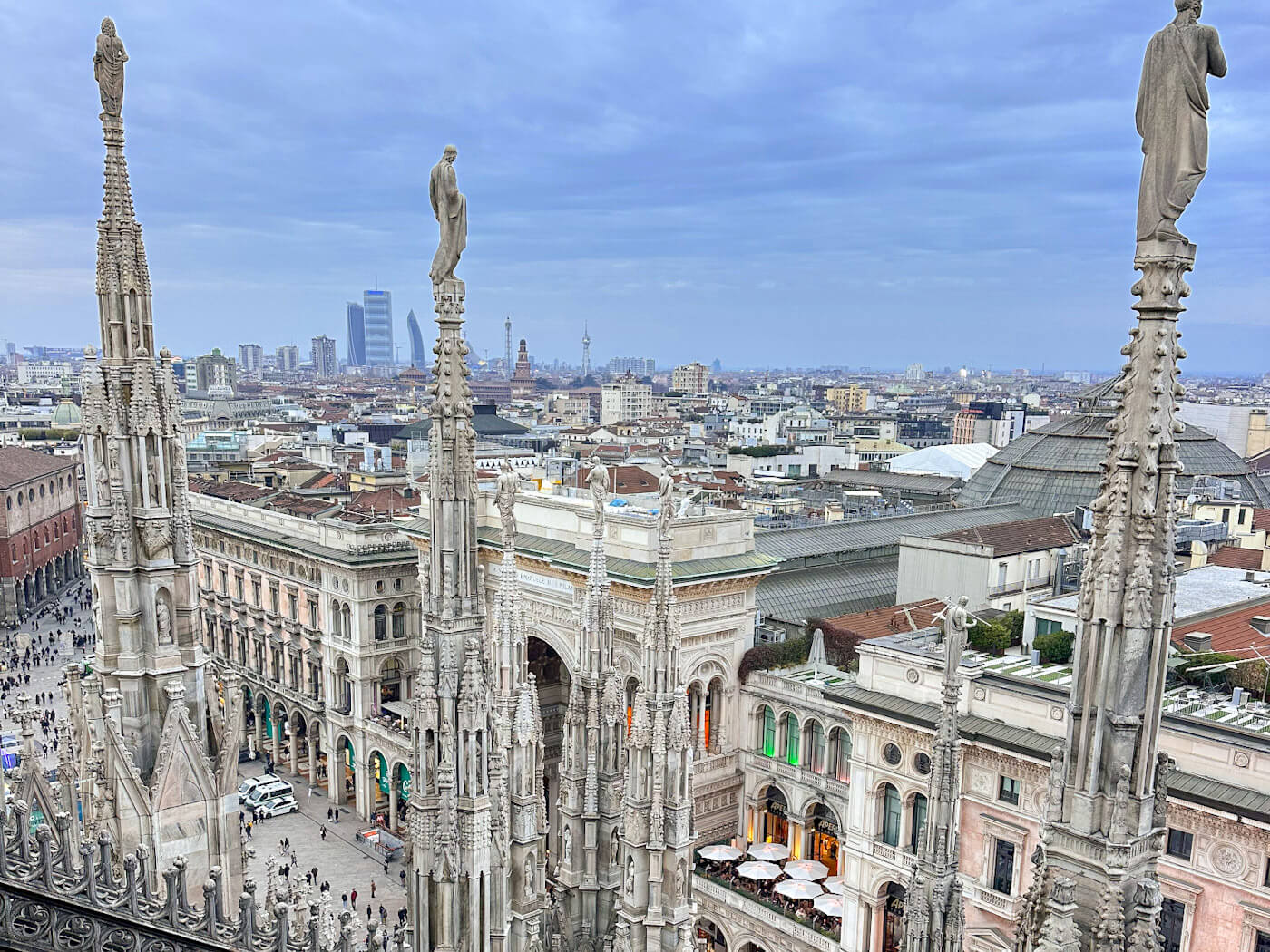 An elevated view from the Milan Cathedral terraces showing intricate Gothic stone spires topped with statues, overlooking the Piazza del Duomo, the Galleria entrance, and the city skyline under a cloudy sky. A must 'what to do in Milan for a day'