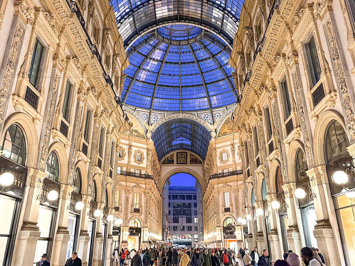 A low-angle shot looking up at the grand glass-domed ceiling of the Galleria Vittorio Emanuele II at dusk, showcasing the ornate neoclassical facades, glowing street lamps, and crowds of people walking through the luxury shopping arcade.