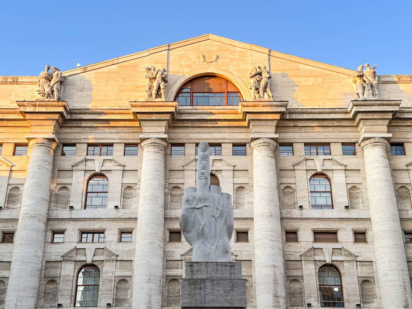 A direct, low-angle view of Maurizio Cattelan’s provocative "L.O.V.E." sculpture, a giant marble hand with all fingers severed except the middle one, positioned in front of the grand, classical facade of the Borsa Italiana (Italian Stock Exchange) building in Milan.
