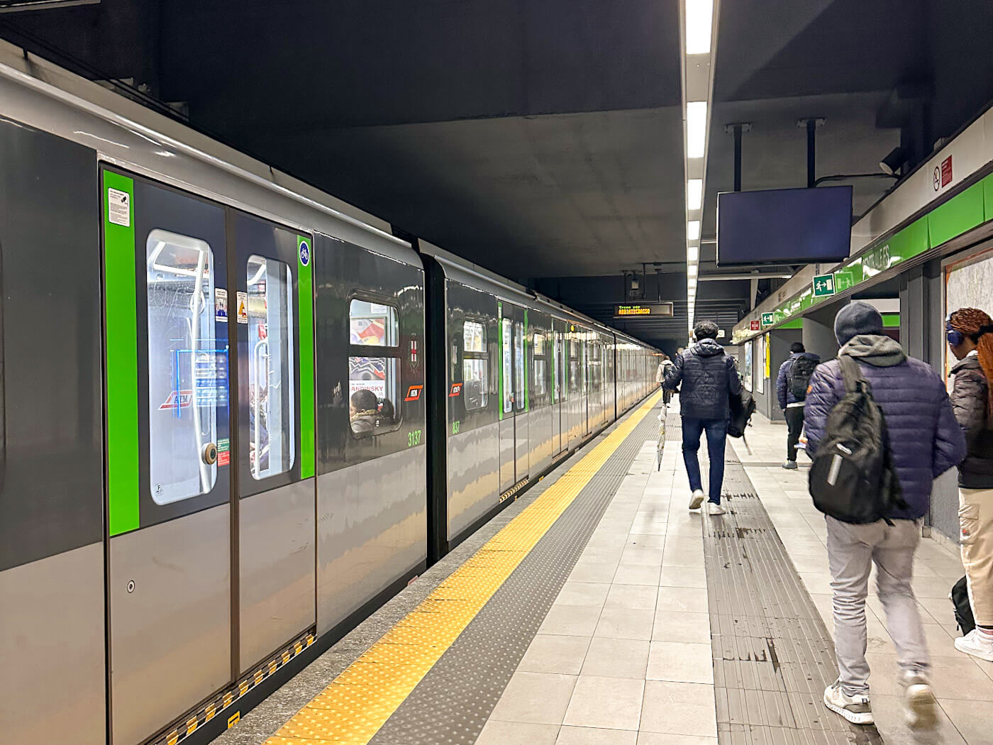 A modern subway platform at the Centrale FS station in Milan, featuring a sleek grey and green train stopped at the platform while commuters with backpacks walk toward the exit under bright overhead lights. How to get around what to do in Milan for a day