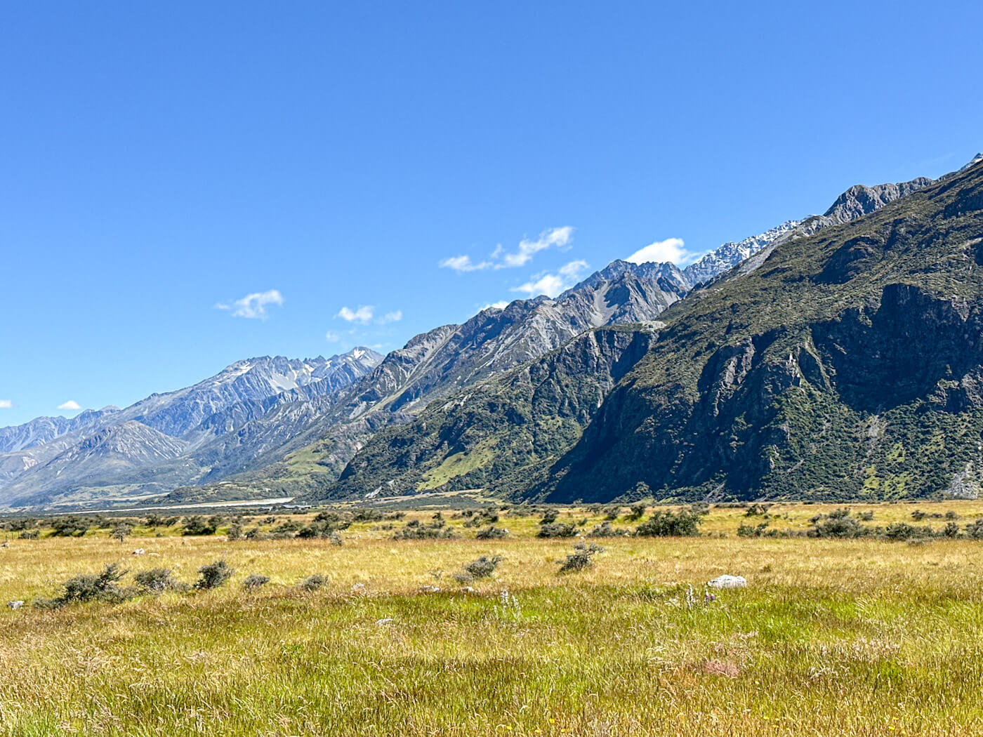A wide landscape view of the vast, golden grasslands of Twizel, New Zealand, set against a backdrop of the Southern Alps under a blue, cloudy sky. This area served as one of the major Lord of the Rings Filming Locations for the Battle of the Pelennor Fields