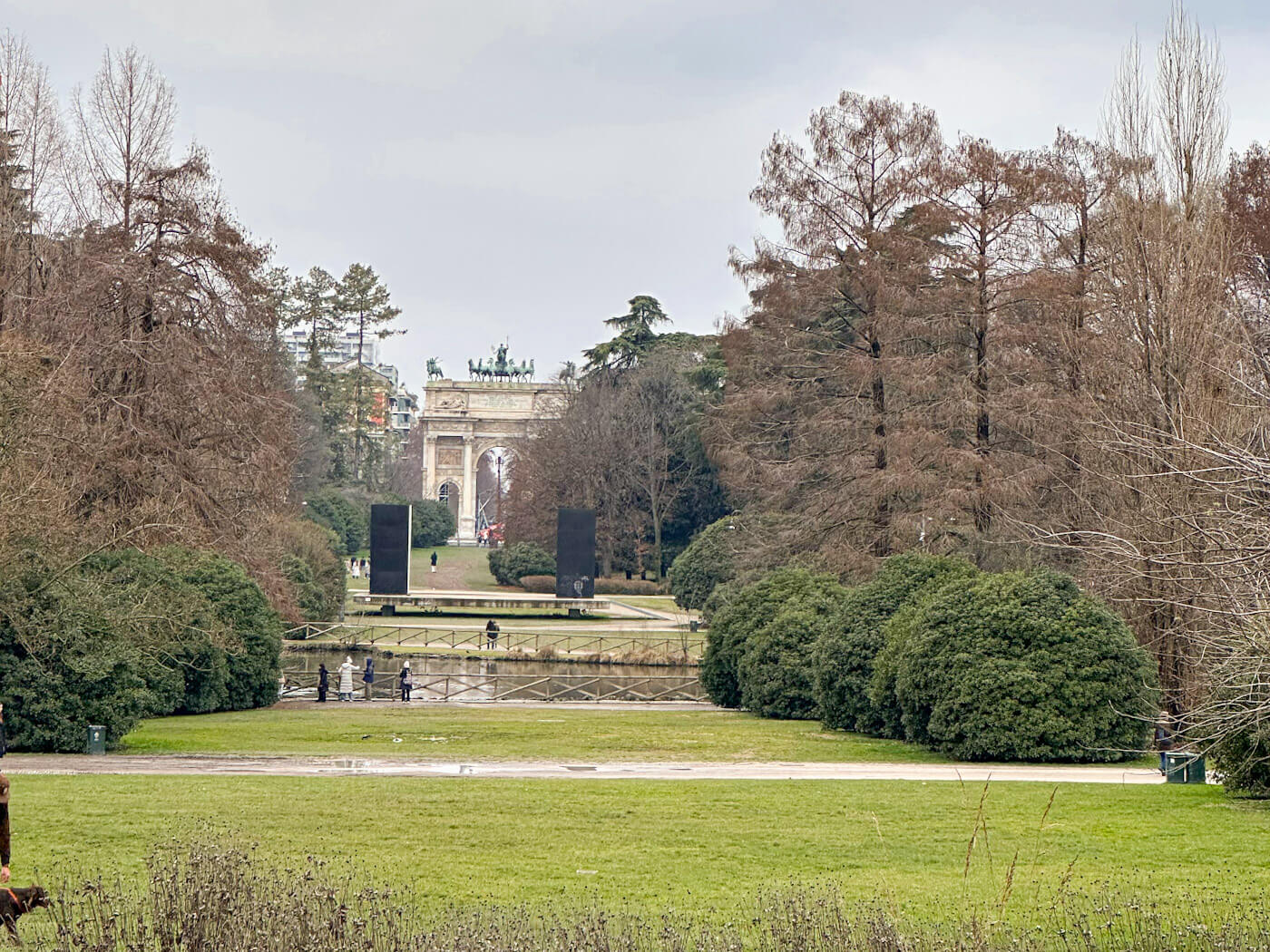 A long-distance view through the lush greenery of Parco Sempione, featuring a green lawn and a small pond in the foreground with the white marble Arco della Pace (Arch of Peace) visible at the end of a tree-lined path.