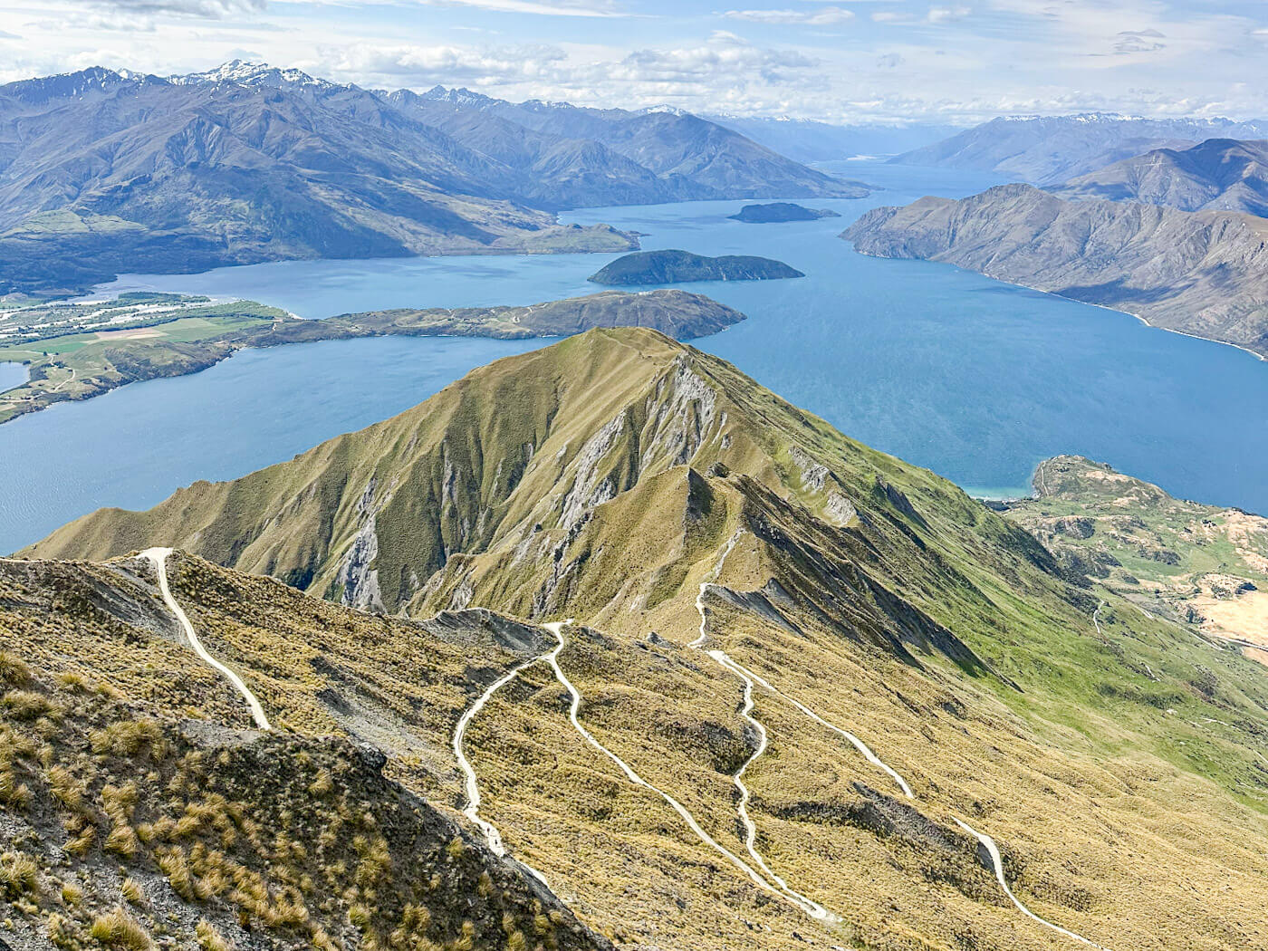 An expansive landscape shot showing the scale of the Roy's Peak track. Multiple zig-zagging switchbacks of a light-colored dirt trail are visible carved into the golden-brown mountainside. The trail leads toward a sharp central ridge, with the vast blue lake and distant snowy mountains filling the rest of the background.