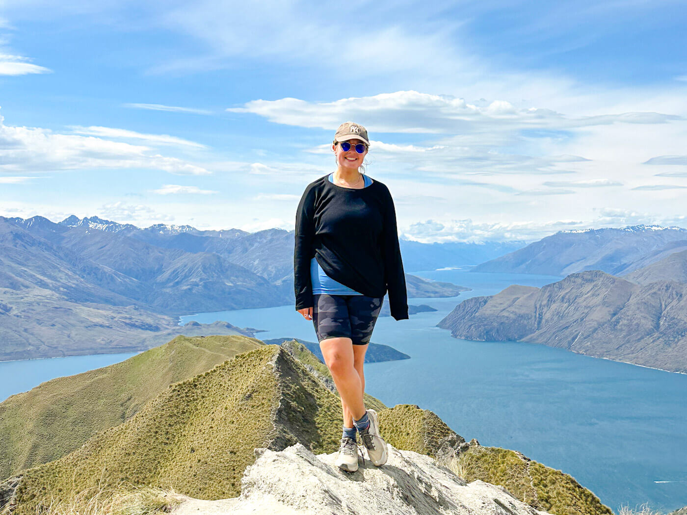 Charlie is smiling on a rocky peak in the foreground, wearing a black long-sleeved shirt, camouflage shorts, a baseball cap, and sunglasses. Behind her, the dramatic scenery of the sharp mountain ridge, Lake Wฤnaka, and the distant snowy Southern Alps unfolds under a clear sky.